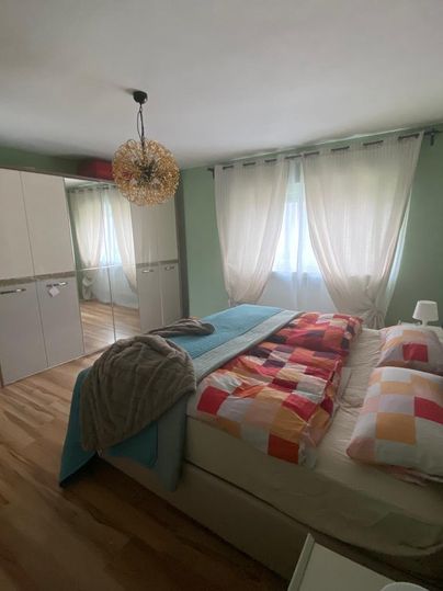 Ein gemütliches Schlafzimmer mit einem großen Bett und bunten Kissen. Der Raum hat helle Vorhänge und einen Spiegel in einem Schrank.