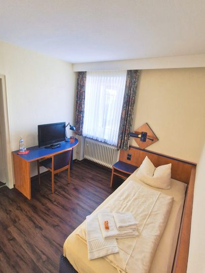 A simple hotel room with a single bed and a desk. There is a television and plenty of natural light coming through the window.