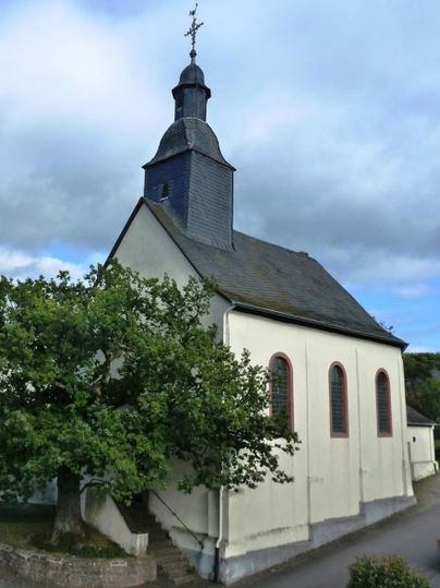 Eine kleine Kirche mit einem grauen Turm und großen Fenstern. Im Vordergrund steht ein grüner Baum.