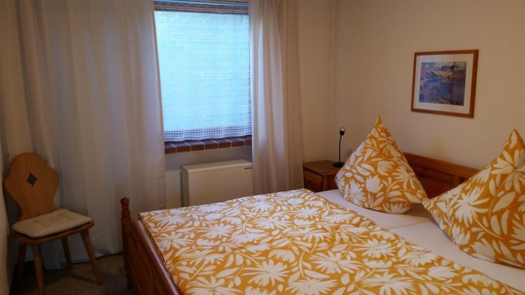 Ein gemütliches Schlafzimmer mit einem Doppelbett, bunten Kissen und einem Fenster, das natürliches Licht hereinlässt. Eine schöne Holzkommode und ein Stuhl ergänzen die Einrichtung.