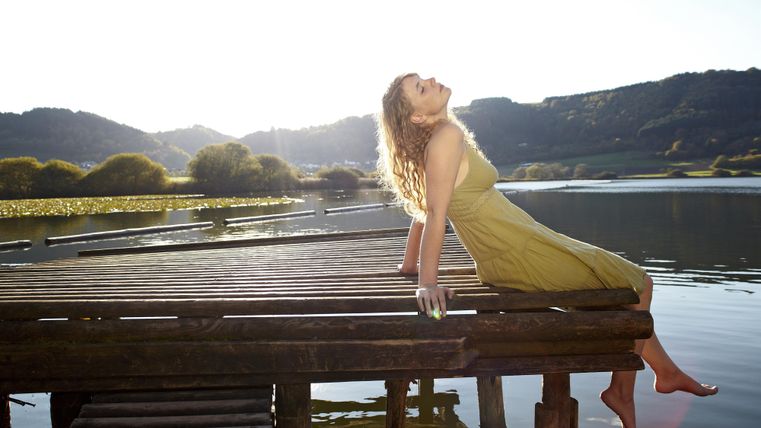 Eine Frau sitzt entspannt auf einem Holzsteg am Wasser. Die Sonne scheint hell und die Umgebung ist malerisch.