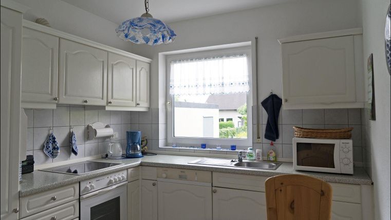 Eine helle Küche mit weißen Schränken und modernen Geräten. Ein Tisch mit einem Stuhl steht in der Mitte, und ein Fenster sorgt für natürliches Licht.