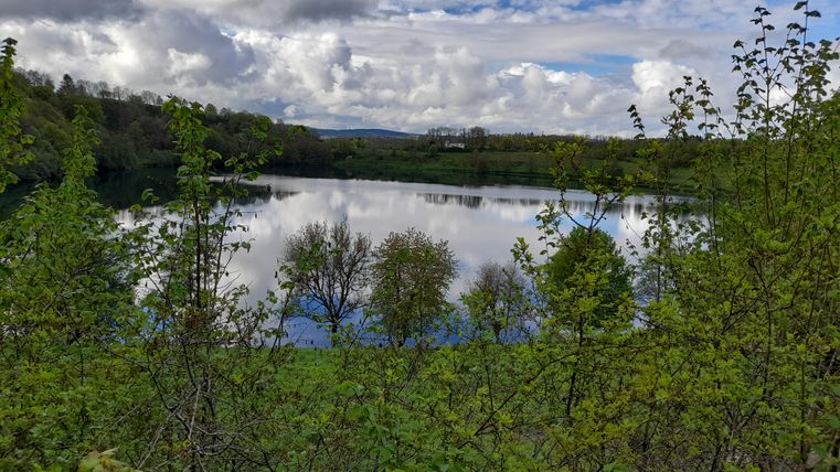 A quiet lake surrounded by green trees and gentle hills. The sky is overcast, but the landscape radiates a peaceful atmosphere.