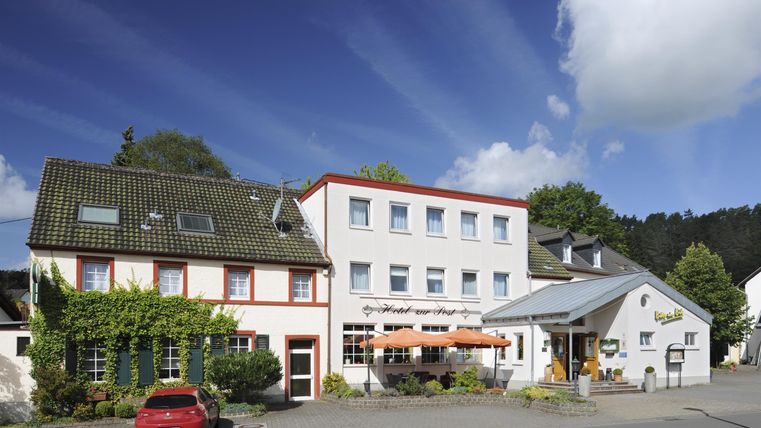 Ein zweigeschossiges Gebäude mit einem angemalten Obergeschoss und einem darunter liegenden Restaurantbereich. Vor dem Gebäude steht ein rotes Auto und die Umgebung ist grün und einladend.