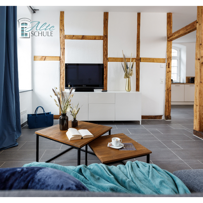 Ein modernes Wohnzimmer mit Holzbalken und einem Fernseher an der Wand. Auf dem Tisch liegen ein Buch und eine Tasse, ergänzt durch eine stilvolle Dekoration.