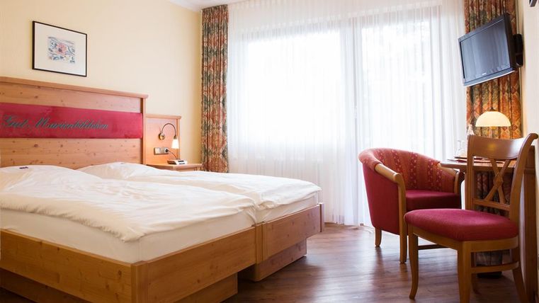 Ein gemütliches Hotelzimmer mit zwei Betten und Holzinterieur. Große Fenster lassen viel Tageslicht herein und es gibt einen kleinen Arbeitsplatz mit Stuhl.