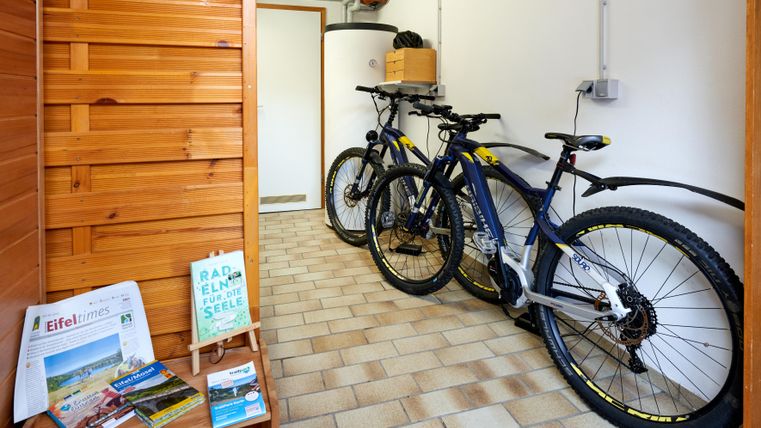 Two bicycles are standing in a small room next to wooden shelves. On a table, there are some brochures and information about the region.