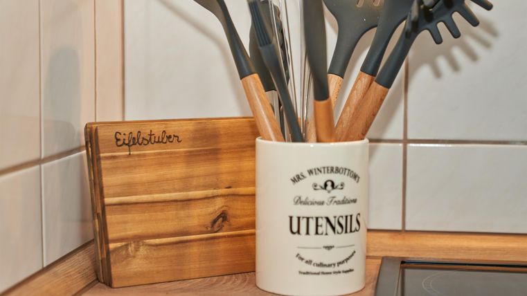A kitchen cabinet with a pot holder and various cooking utensils. The container is white with an inscription, and the utensils have wooden elements.