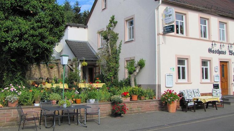 Ein charmantes Gasthaus mit blühendem Garten und gemütlichen Sitzplätzen im Freien. Die Fassade ist freundlich und einladend gestaltet.