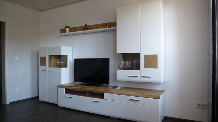 Ein modernes Wohnzimmer mit einem weißen Fernsehmöbel und einer großen TV-Anzeige. Die Wand ist hell und schlicht gestaltet, mit minimalistischem Stauraum.