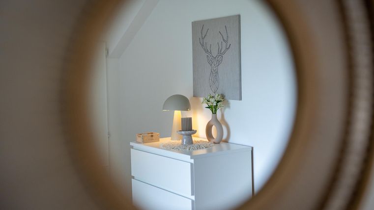 Ein helles und helles Zimmer mit einem weißen Kommode, einer Lampe und einer dekorativen Pflanze. An der Wand hängt ein Bild mit einem Hirschmotiv.