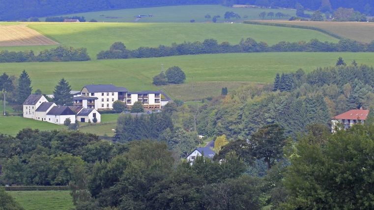 A picturesque landscape with green meadows and gentle hills. In the foreground, some houses and trees are visible.