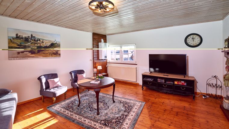 A cozy living room with wooden flooring and a bright ceiling. There is a table, two chairs, and a television on the wall.