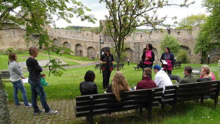 Eine Gruppe von Wanderen mit ihrer Krimiführerin, teils auf Bänken sitzend, teils stehend vor der Kulisse der Stadtmauer in Hillesheim