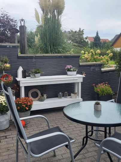 A cozy outdoor area with a table and chairs. Many plants and flowers decorate the surroundings.