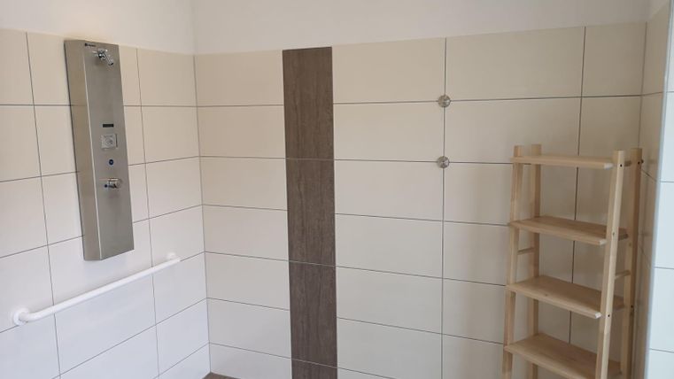 A bright shower area with white tiles and a wooden shelf. A shower fixture and grab bars are mounted on the wall.