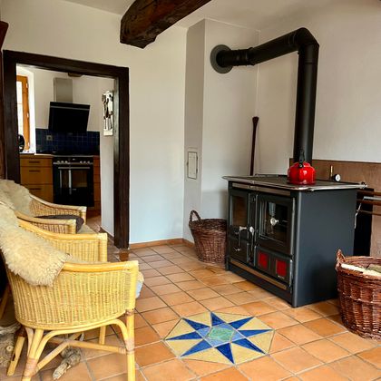 Ein gemütlicher Raum mit einem Holzofen und zwei rattan Stühlen. Der Boden ist mit gefliesten Farben verziert und es gibt einen Blick auf eine angrenzende Küche.