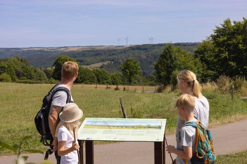Eine Familie steht vor einem Informationsschild in einer grünen Landschaft. Im Hintergrund sieht man hügelige Felder und Bäume unter einem klaren Himmel.