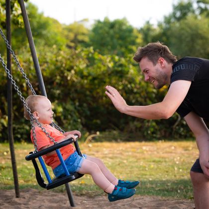 A smiling baby is crawling on a sofa while two adults and another toddler watch in the background. The atmosphere is friendly and cheerful.