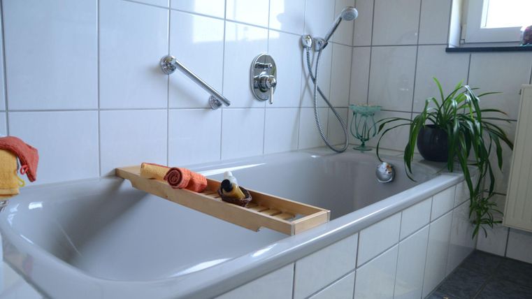 Een lichte badkamer met een bad en witte tegels. Op het bad ligt een houten dienblad met badelementen en een plant staat op de achtergrond.
