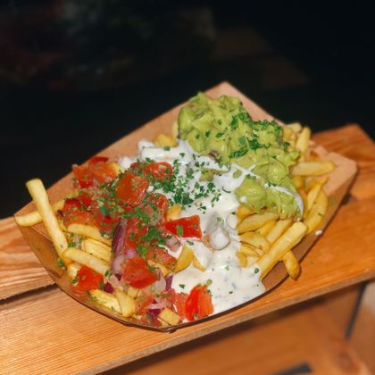 Pommes in einer Holzschale, garniert mit Guacamole, Tomatenstücken und weißer Soße, dekoriert mit Kräutern.