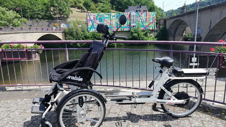 Ein Fahrrad steht an einem Flussufer, umgeben von grüner Landschaft. Im Hintergrund sind bunte Wandmalereien und eine Brücke zu sehen.