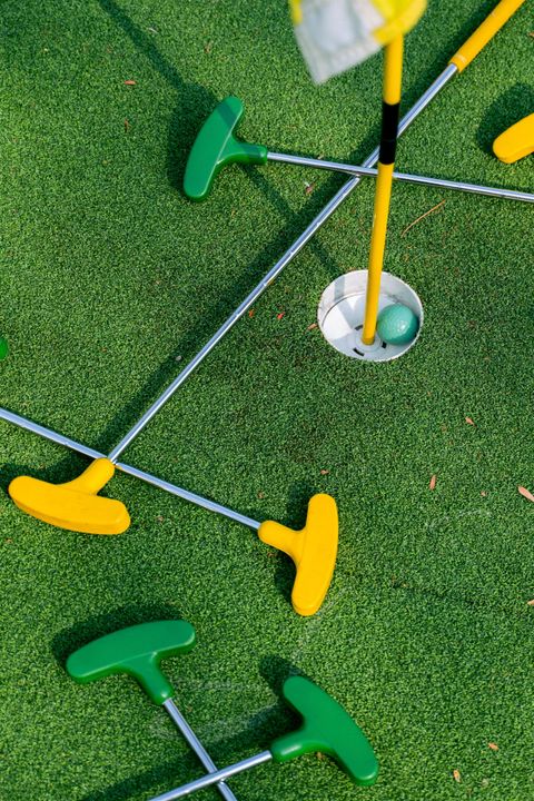 In der Mitte des Bildes sieht man ein Loch, indem ein grüner Golfball liegt. Um das Loch liegt grüner Kunstrasen, auf dem mehrere Schläger liegen. Die Schläger haben gelbe und grüne Schlägerköpfe, der Schaft ist metallfarbend.