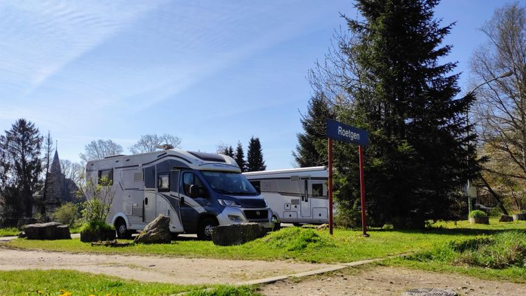 A beautiful campsite with two motorhomes and a green environment. In the background, trees and a path are visible.