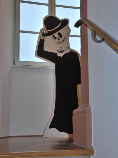 Eine stylisierte Figur in Schwarz trägt einen Hut und steht an einer Treppe. Der Hintergrund zeigt ein Fenster mit natürlichem Licht.