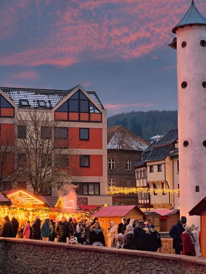 Een feestelijke kerstmarkt met verlichte kraampjes en veel bezoekers. De lucht is rood-oranje gekleurd en de gebouwen op de achtergrond zijn winterlijk versierd.