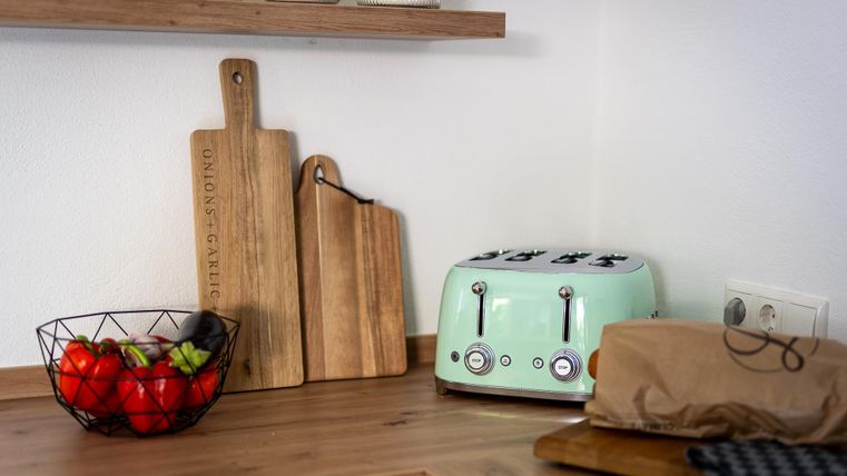 Eine moderne Küche mit einem grünen Toaster und frischem Gemüse in einer Schüssel. Holzbrettchen liegen dekorativ auf der Arbeitsplatte.