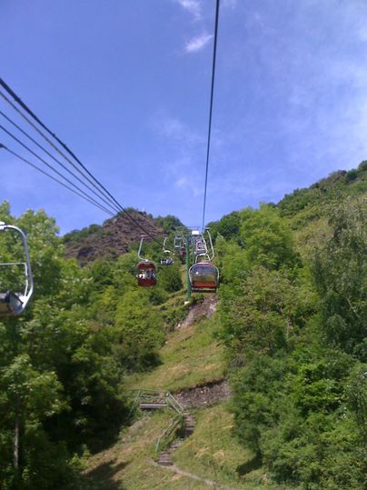 Een kabelbaan rijdt door een groene landschap. Op de achtergrond zijn er bomen en een blauwe lucht te zien.