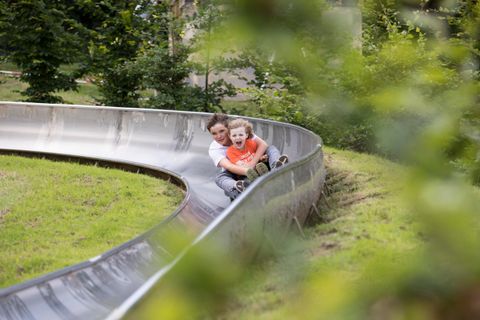 Zwei Kinder fahren gemeinsam eine Rodelbahn durch einen grünen Park. Sie lachen und genießen die Fahrt.