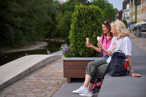 Two women are sitting on a bench by the water, enjoying ice cream. In the background, trees and colorful houses can be seen.