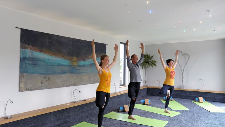 Drei Personen praktizieren Yoga in einem hellen Raum. Sie stehen auf grünen Matten und halten ihre Arme erhoben.