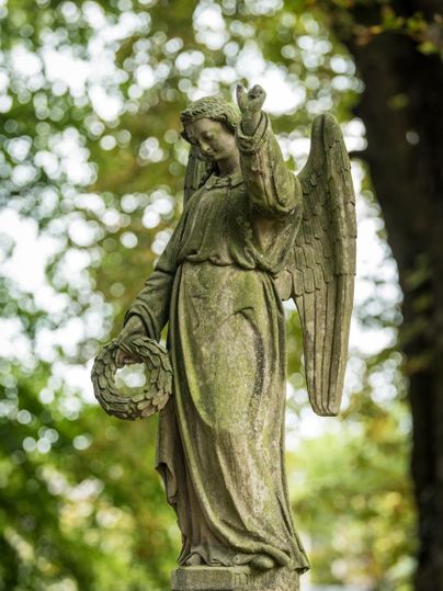 Een standbeeld van een engel met vleugels en een krans in de hand. Het standbeeld staat omringd door groene bomen.
