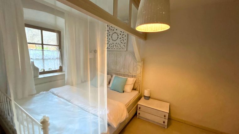 A cozy bedroom with a bed surrounded by a white curtain. A nightstand and a warmly lit ceiling lamp create an inviting atmosphere in the room.
