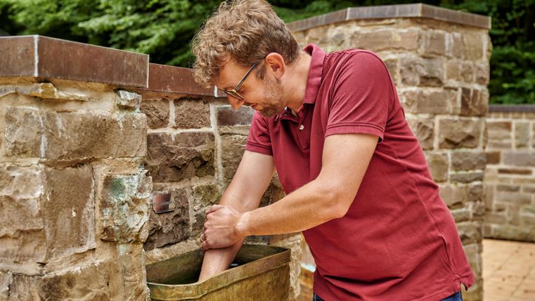 Een man staat naast een stenen muur en wast zijn handen in een wasbak. De omgeving is groen en natuur dichtbij.