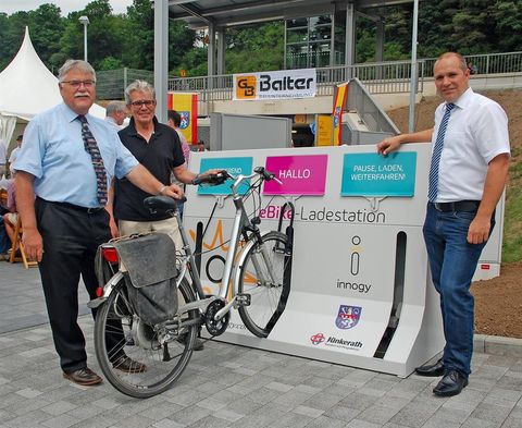 Three men are standing next to an e-bike charging station. A bicycle is connected to the station. In the background, a tent and a building can be seen.