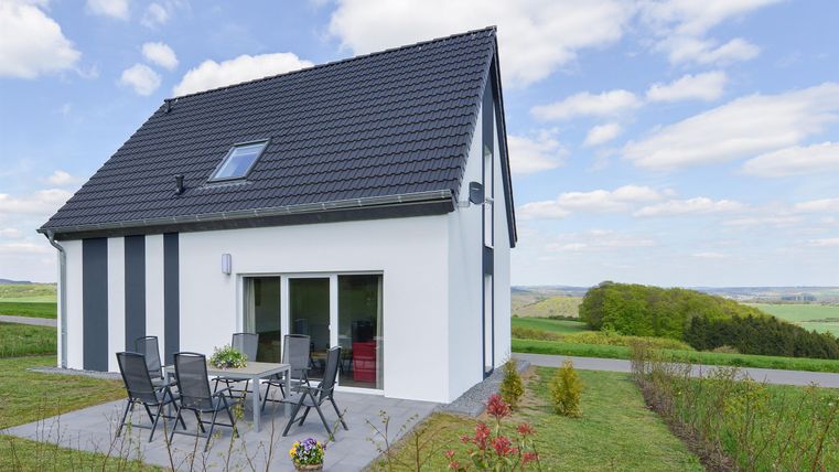 Ein modernes Haus mit schwarzem Dach und weißen Wänden. Es gibt eine Terrasse mit Stühlen und einen weiten Blick auf die grüne Landschaft.