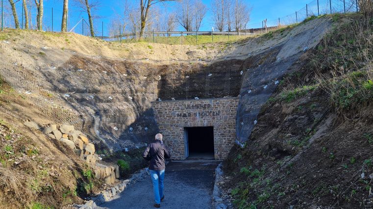 Een steile helling met een ingang naar een historisch bouwwerk. Op de voorgrond loopt een persoon op het pad naar de opening.