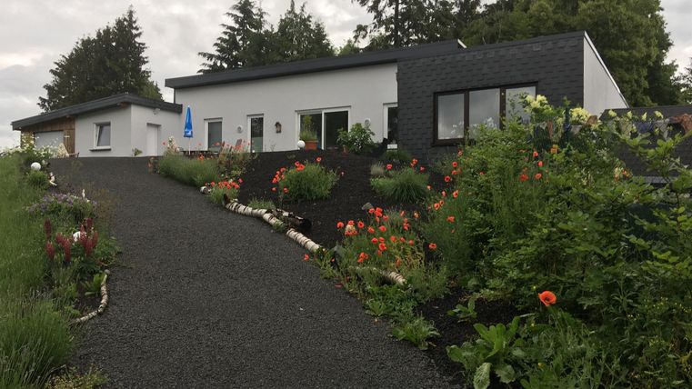 A modern house with a well-maintained garden and colorful flowers. The path to the house is covered with gravel and surrounded by trees.