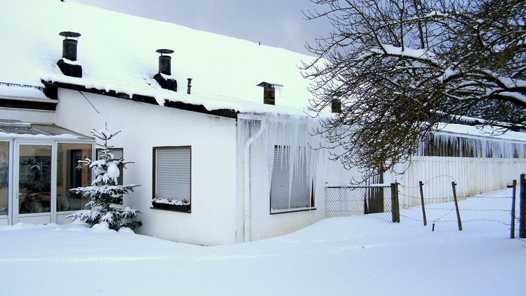 Ein Haus im Winter, bedeckt von Schnee mit Eiszapfen an der Dachkante. Die Umgebung ist ruhig und winterlich.