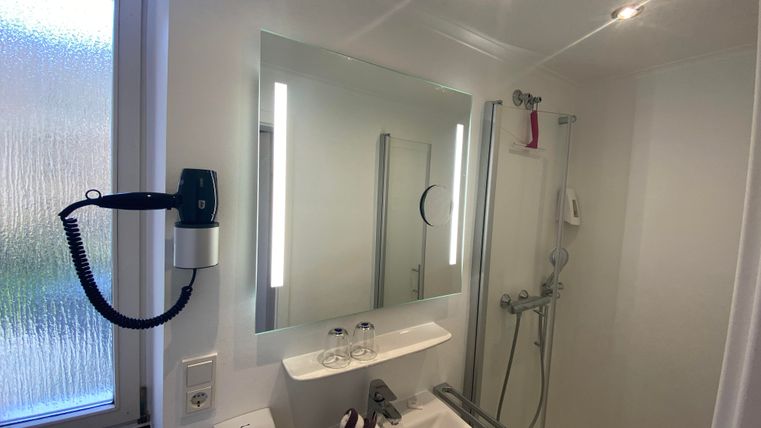 Eine moderne Badezimmeransicht mit einem Spiegel, einer Duschkabine und einem Waschbecken. Die Wände sind hell und es gibt große Fenster, die Licht hereinlassen.