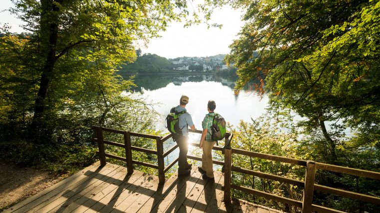 Zwei Personen stehen auf einem Holzsteg und blicken auf einen See, umgeben von Bäumen.