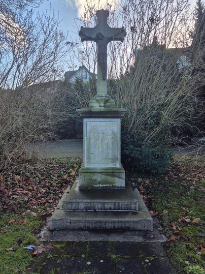An old gravestone with a cross stands in a grassy area. Surrounded by dry branches and grass, the scene is calm and contemplative.