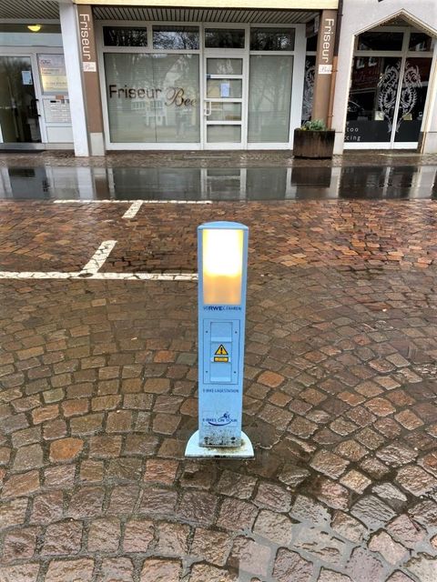 A blue light pole stands on a wet, paved sidewalk. In the background, shops can be seen.