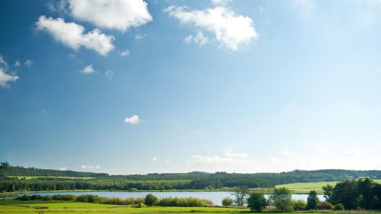 Eine malerische Landschaft mit einem ruhigen See, umgeben von grünen Wiesen und Bäumen. Der Himmel ist klar und sonnig mit einigen wenigen Wolken.