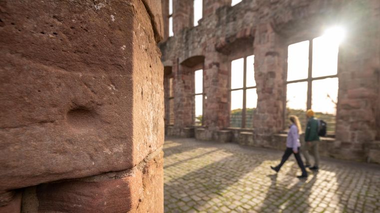 Eine historische Ruine mit großen Fenstern und Sonnenlicht, das hereinfällt. Zwei Personen gehen entspannt über den steinernen Boden.
