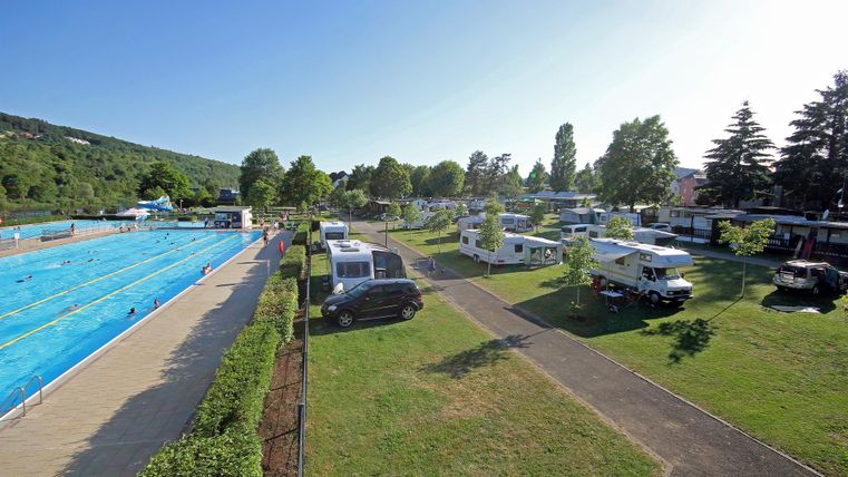 An outdoor swimming pool with blue water basins surrounded by many trees. In the background, motorhomes and a green campsite can be seen.